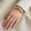 Hand wearing polished gold wavy cuff bangle with white sweater - Avenzra Gold Wavy Bangle
