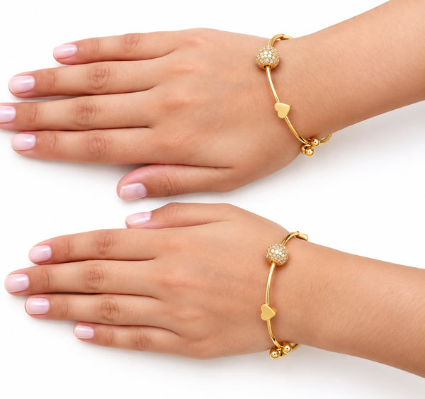 Two hands wearing pair of gold charm bangles with heart, flower and stone ball - Avenzra Gold Charm Bangle